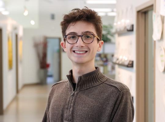 A young man with glasses and a brown quarter-zip sweater smiling at the camera.